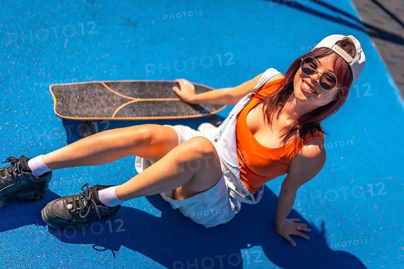 Top view of a cool urban woman with sunglasses and cap sitting with her skateboard in a colorful park