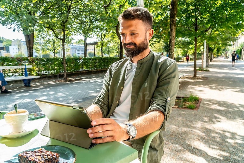 Bearded man at an outdoor cafe working on a tablet, enjoying coffee and cake in a sunny parklike setting while browsing and staying connected to work remotely