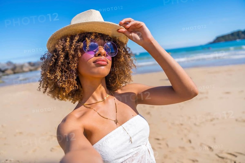 Personal perspective of a beauty latin young woman wearing sunglasses and sun hat taking a selfie enjoying summer at sandy beach