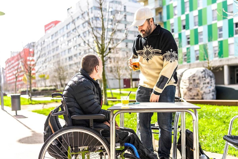 Two friends enjoying drinks at an outdoor table, one using a wheelchair, demonstrating inclusion and friendship in a casual setting