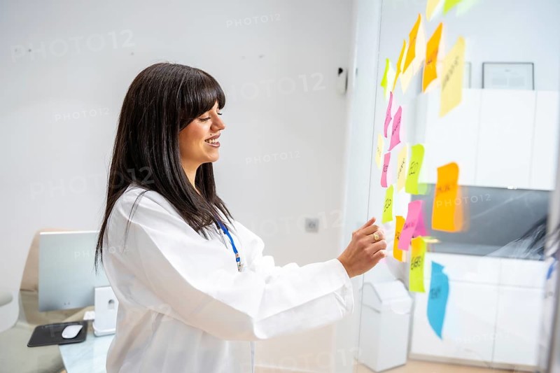 Female doctor in a white coat brainstorming and planning healthcare strategies, organizing ideas with colorful sticky notes on a transparent glass board in a modern clinic or hospital