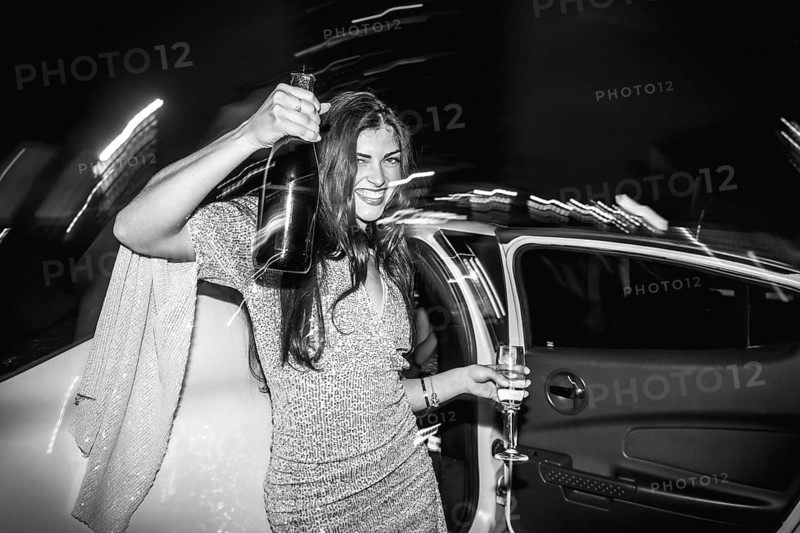 Happy young woman exiting a luxury limousine car, holding a champagne bottle and glass, celebrating a night out at a party, depicted in black and white with motion blur