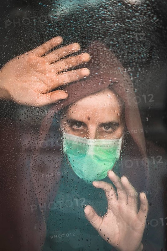 Sad look of a young man with a mask in the Covid-19 pandemic looking through a window