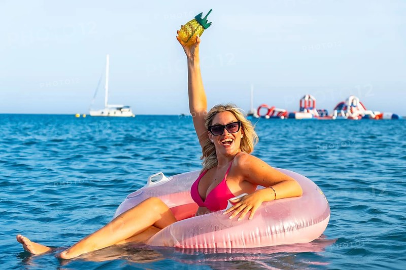 Blonde woman celebrating her summer vacation, sitting on a pink inflatable ring while holding a tropical drink with her arm raised, smiling and having fun in the blue sea