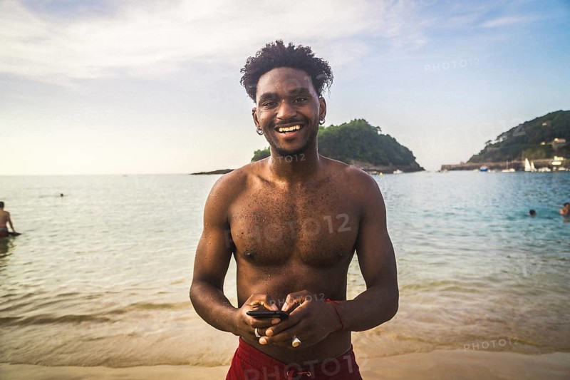 Young man standing in shallow ocean water on a sandy beach, holding a smartphone and smiling, enjoying a casual moment on a warm summer day in a retro style