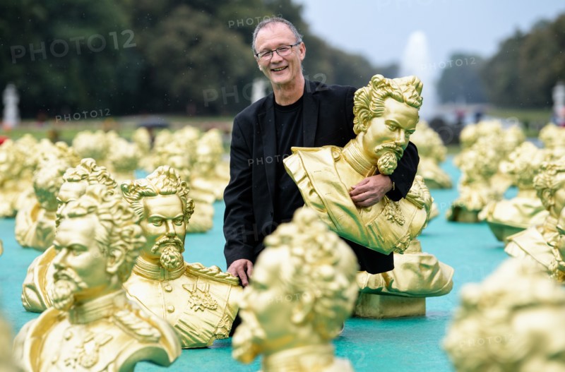 24.08.2018, Bayern, München: Der Nürnberger Künstler Ottmar Hörl blickt neben seiner Kunstinstallation zu Ehren des Märchenkönigs König Ludwig II. im Nymphenburger Schlosspark in die Kamera. 100 Abbilder Ludwigs II. wurden im Rahmen der Kunstinstallation einen Tag vor Ludwigs 173. Geburtstag aufgestellt. Foto: Matthias Balk/dpa | Verwendung weltweit