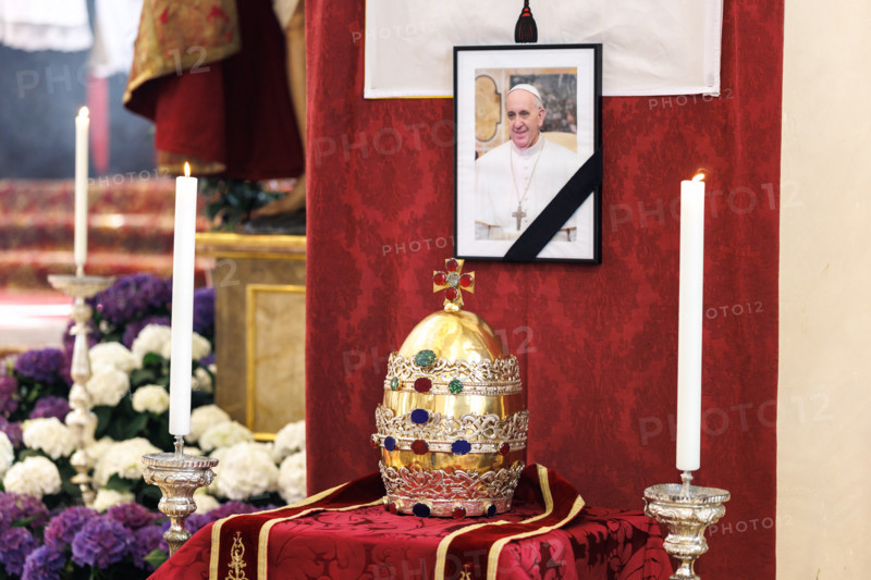 Eine Papstkrone, die sogenannte Tiara, liegt vor einem Bild vom verstorbenen Papst. Zu Ehren des gestorbenen Pontifex wurde der Figur vom Kirchenpatron Petrus am Hauptaltar der Kirche die Papstkrone abgenommen. Dieses Ritual wird immer dann ausgeführt, wenn ein Papst stirbt.