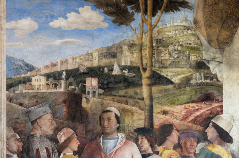 Ducal Palace: south-west wall of the “Bridal Chamber or Camera Picta", which is a room located in the north-east tower of the Castle of San Giorgio in Mantua. It is famous for the cycle of frescoes that covers its walls, a masterpiece by Andrea Mantegna, created between 1465 and 1474.
The wall, on the theme of “The Meeting” is divided into three sectors. In the one on the left the actual meeting takes place, in the central one some cherubs hold a dedicatory plaque and in the one on the right the marquis's court parades.
Detail of the right part of the wall.
The Marquis Ludovico is represented in the Meeting, this time in official guise, perhaps flanked by Ugolotto Gonzaga, son of his deceased brother Carlo. His son Francesco who is a Cardinal is in front of him. Below them are the sons of Federico I Gonzaga, Francesco and Sigismondo, while their father Federico is on the far right: the generous folds of his dress are a ploy to hide his kyphosis. Federico is in conversation with two ...