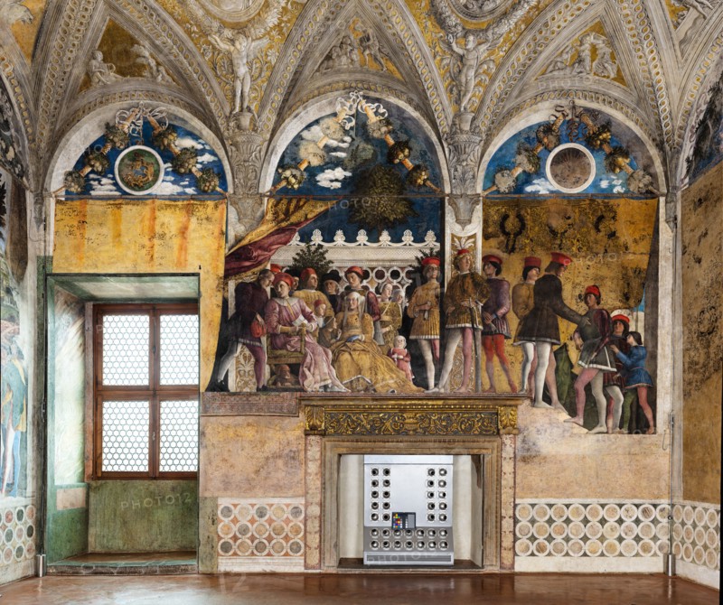 Ducal Palace: north-west wall of the “Bridal Chamber or Camera Picta", which is a room located in the north-east tower of the Castle of San Giorgio in Mantua. It is famous for the cycle of frescoes that covers its walls, a masterpiece by Andrea Mantegna, created between 1465 and 1474.
This wall represents “The Court” of Ludovico II Gonzaga. On the left, under the opened curtain, Ludovico sits while a secretary (perhaps Marsilio Andreasi or Raimondo Lupi di Soragna) hands him a letter. We see Rubino, Marquis Ludovico's favorite dog and symbol of loyalty, crouching. To her right, the only other person seated, is the Marquis's wife, Barbara of Brandenburg with a little girl, perhaps the youngest daughter Paola who seems to be offering her an apple. Behind Ludovico we can recognize the third-born Gianfrancesco standing, holding his hands on the shoulders of a child, perhaps the prothonotary Ludovicino. The man with the black hat is Vittorino da Feltre, tutor of the marquis and his childre...