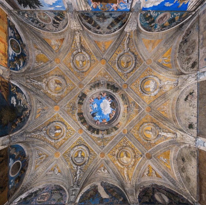Ducal Palace: vault of the “Bridal Chamber or Camera Picta", which is a room located in the north-east tower of the Castle of San Giorgio in Mantua. It is famous for the cycle of frescoes that covers its walls, a masterpiece by Andrea Mantegna, created between 1465 and 1474.
Detail of the vault illusionistically divided into painted rib vault and pendentives. Fake ribs divide the space into regular figures, with a golden background and monochrome paintings.
In the center is the famous oculus. It is a round open illusionistically towards the sky, which was supposed to recall the famous oculus of the Pantheon. In the oculus you can see a balustrade from which a court lady leans out, accompanied by her black servant, a group of maids, a dozen putti, a peacock and a vase, against the background of a blue sky. To reinforce the impression of the open oculus, Mantegna painted some cherubs dangerously poised clinging to the inside of the frame, with dizzying glimpses of their plump little bo...