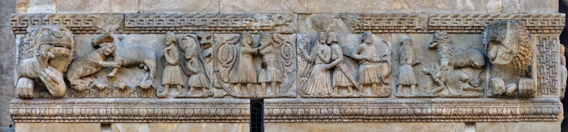 Fidenza, duomo (cattedrale di San Donnino), torre di destra (Torre del Trabucco), fronte ovest: fregio composto da cinque formelle delimitate da due grandi teste di leone. La tesi oggi più accreditata vi individua le “Storie di Berta, Milone e Rolandino” raccontate da due chanson de geste tra la fine del XII e l'inizio del XIII secolo. 
La lettura delle formelle deve essere condotta da destra a sinistra, individuando le seguenti scene:
Pipino il Breve salva a mani nudi due "lombardi" dalle fauci di un leone;
Carlo Magno va a caccia, mentre Milone seduce Berta, sorella di Carlo Magno;
Milone lotta con un bandito che vorrebbe rapire Berta rimasta incinta;
Milone, seguito dal figlio Rolandino, va nel bosco per procurarsi legna da vendere;
Un leoncino – allegoria di Rolandino – azzanna al collo un cavallo.
Opera di Benedetto Antelami e bottega.


[ENG]
Fidenza, duomo (Cathedral of San Donnino), right tower (Trabucco Tower), west front: frieze composed of five panels delimited by...