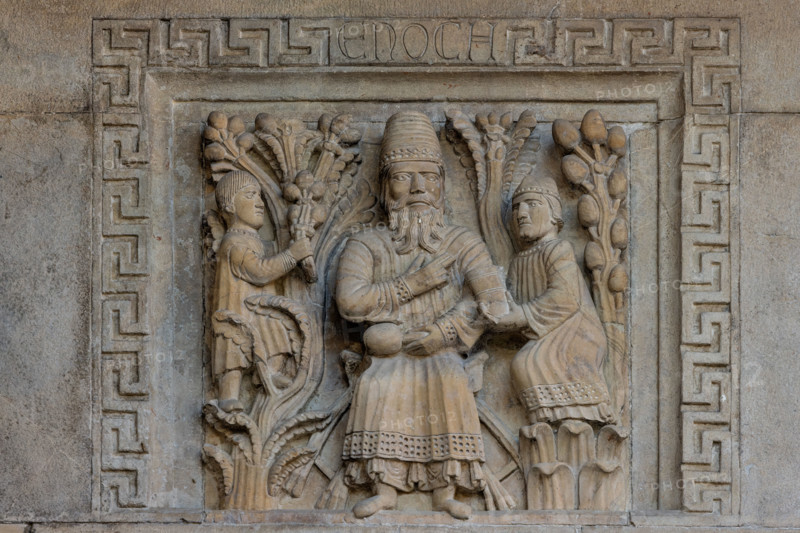 Fidenza, Duomo (Cattedrale di S. Donnino), facciata: bassorilievo con "Il profeta Enoch” di Benedetto Antelami e bottega.
Il personaggio è assiso su un faldistorio nel mezzo d’un giardino (Il Paradiso) rappresentato da due alberi carichi di frutti. È vestito con una tunica e un soprabito più corto con orli lavorati, e porta in testa un cappello troncoconico. Benedice con la destra, e nella sinistra tiene un oggetto che sembra un frutto. Ha ai fianchi due assistenti; quello a destra gli porge un contenitore cilindrico, mentre quello a sinistra è arrampicato su un albero intento a cogliere dei frutti. 

[ENG]
Fidenza, Duomo (Cathedral of S. Donnino), façade: bas-relief with "The Prophet Enoch" by Benedetto Antelami and workshop.
The figure is seated on a faldstool in the middle of a garden (Paradise) represented by two trees laden with fruit. He is dressed in a tunic and a shorter overcoat with worked edges, and wears a truncated cone-shaped hat. He blesses with his right hand, and ...