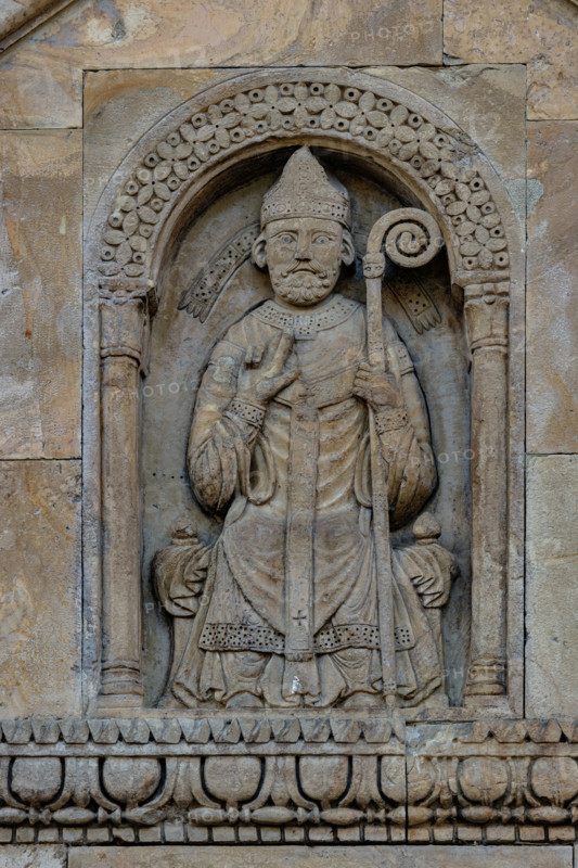 Fidenza, Duomo (Cattedrale di San Donnino), facciata, timpano del portale destro: “figura di Vescovo”. il personaggio è seduto su un faldistorio, vestito di camice, cotta e casula, con mitria e pastorale. Tradizionalmente nella figura è stato riconosciuto l’Arciprete della chiesa di S. Donnino, ma il pallio riservato sin dal medioevo agli arcivescovi metropoliti e ai primati, fa dubitare di questa interpretazione. Opera di Benedetto Antelami e bottega.

[ENG]
Fidenza, Duomo (St. Donnino Cathedral), the facade, tympanum of the right portal: “figure of a Bishop”. The figure is seated on a faldstool, dressed in a cassock, surplice and chasuble, with a mitre and pastoral staff. Traditionally, the figure has been identified as the Archpriest of the church of S. Donnino, but the pallium reserved since the Middle Ages for metropolitan archbishops and primates casts doubt on this interpretation. Work by Benedetto Antelami and workshop.
Cattedrale di San Donnino, Fidenza (PR), Emilia-Romagn...