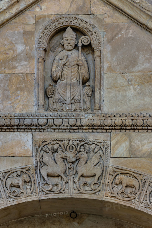 Fidenza, Duomo (Cattedrale di San Donnino), facciata, timpano del portale destro: “figura di Vescovo”. il personaggio è seduto su un faldistorio, vestito di camice, cotta e casula, con mitria e pastorale. Tradizionalmente nella figura è stato riconosciuto l’Arciprete della chiesa di S. Donnino, ma il pallio riservato sin dal medioevo agli arcivescovi metropoliti e ai primati, fa dubitare di questa interpretazione. Opera di Benedetto Antelami e bottega.

[ENG]
Fidenza, Duomo (St. Donnino Cathedral), the facade, tympanum of the right portal: “figure of a Bishop”. The figure is seated on a faldstool, dressed in a cassock, surplice and chasuble, with a mitre and pastoral staff. Traditionally, the figure has been identified as the Archpriest of the church of S. Donnino, but the pallium reserved since the Middle Ages for metropolitan archbishops and primates casts doubt on this interpretation. Work by Benedetto Antelami and workshop.
Cattedrale di San Donnino, Fidenza (PR), Emilia-Romagn...