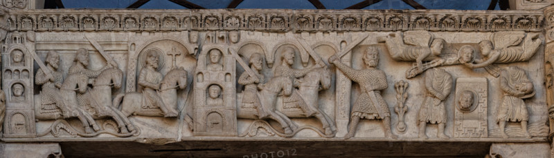Fidenza, duomo (Cattedrale di San Donnino), facciata, portale maggiore: dettaglio dell’archivolto con “Storie di San Donnino”.
Donnino prosegue la fuga verso Piacenza che gli chiude le porte, poi verso un'altra città finché gli sgherri a cavallo lo raggiungono.
Un soldato mozza con la spada la testa del Santo; due angeli la sollevano e la portano in cielo (senza barba); la testa terrena ricompare (con la barba) su una specie di ara; il Santo la prende tra le mani e s'incammina per attraversare il torrente Stirone.
Opera di  Benedetto Antelami e bottega.

[ENG]
Fidenza, duomo (Cathedral of San Donnino), facade, main portal: detail of the archivolt with “Storiee of St. Donnino”.
Donnino prosegue la fuga verso Piacenza che gli chiude le porte, poi verso un'altra città finché gli sgherri a cavallo lo raggiungono;
Un soldato mozza con la spada la testa del Santo; due angeli la sollevano e la portano in cielo (senza barba); la testa terrena ricompare (con la barba) su una specie di a...