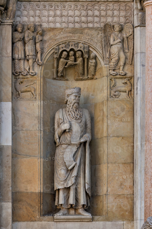 Fidenza, duomo (Cattedrale di San Donnino), sculture tra il portale maggiore e quello di sinistra: la nicchia con il “Re David” di Benedetto Antelami.
Il profeta sorregge un cartiglio sul quale si legge la seguente iscrizione a lettere capitali: DAUID / PPHA / REX / HEC / PORTA / DOMINI / IUSTI: / INTRANT PEREAM (David Propheta Rex – Hec porta Domini iusti intrant per eam [Salmo 118 (117),20]).

Nella calotta della nicchia è la Presentazione di Gesù al Tempio. 
Ai lati sono due formelle con le figura di un grifo e di un capricorno; al di sopra di esse si osserva un bassorilievo raffigurante verosimilmente un Angelo che mostra la via per Roma ad una famiglia di pellegrini ricchi. 

[ENG]
Fidenza, duomo (Cathedral of San Donnino), sculptures between the main portal and the left one: the niche with “King David” by Benedetto Antelami.
The prophet holds a scroll on which the following inscription can be read in capital letters: DAUID / PPHA / REX / HEC / PORTA / DOMINI / IUSTI: / IN...