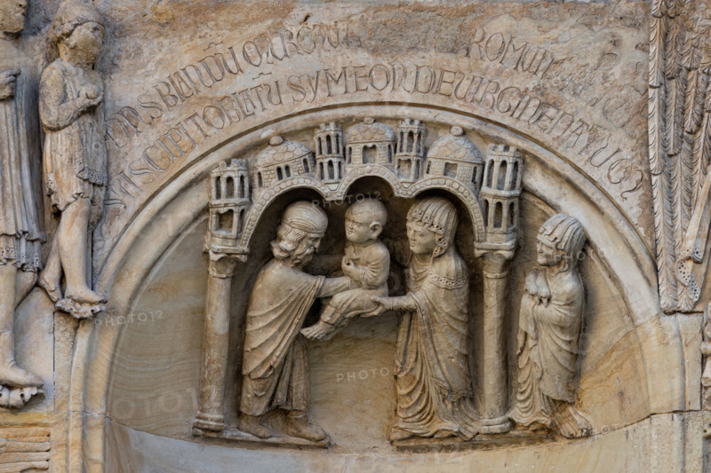 Fidenza, duomo (Cattedrale di San Donnino), sculture tra il portale maggiore e quello di sinistra vicino alla nicchia con il “Re David” di Benedetto Antelami.
Particolare della presentazione di Gesù al tempio di Gerusalemme come stabilito dalla legge di Mosè sul primogenito (Lc 2,22-38); è rappresentata sotto un edificio, delimitato da due colonne con capitelli che sostengono un triplice arco sormontato da cupole e torri, ove Maria offre il Bambino Gesù al sacerdote Simeone, il quale ricevendolo, ringrazia Dio. A destra, fuori dall’edificio, una donna ha in mano le due tortore che, come stabilito dalla legge ebraica, erano offerte in sacrifico in quell’occasione.
Sopra la nicchia è incisa la seguente iscrizione a lettere capitali: DANSBLANDU MURMU[RDATURP]ROMUN[E]RETUR[TUR] / SUSCIPITOBLATU SYMEONDEVIRGINENATUM (Dans blandum murmur datur pro munere turtur. / Suscipit oblatum Simeon de Virgine natum).

[ENG]
Fidenza, duomo (Cathedral of San Donnino), sculptures between the main por...