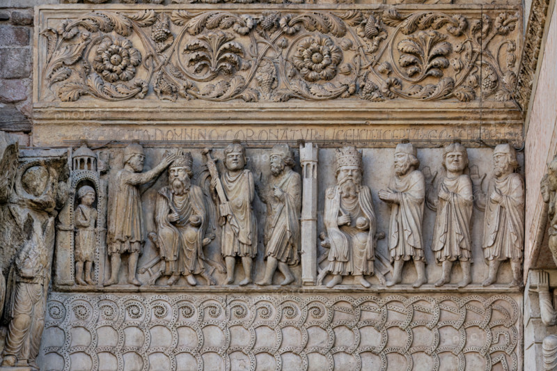 Fidenza, duomo (Cattedrale di San Donnino), sculture tra il portale maggiore e quello di sinistra vicino alla nicchia con il “Re David” di Benedetto Antelami.“Storie di San Donnino” con il primo e secondo episodio: Nel pannello a sinistra, la scena è rappresentata all’esterno d’un edificio sotto il quale si trova una guardia armata. 
Donnino, cubiculario di Massimiano Erculeo, stando alla destra dell’imperatore, gli pone la corona sul capo; alla sinistra di Massimiano il Portaspada dimostra che la scena costituisce un momento dell’esercizio del potere sovrano di giurisdizione, probabilmente nei confronti del personaggio posto all’estrema destra. Nella cornice attigua posta sopra è incisa a lettere capitali l’iscrizione: IR . ABEATO . DOMNINO CORONATVR (Imperator a Beato Domnino coronatur = L’Imperatore è incoronato dal beato Donnino).
Nel pannello a destra la distinzione dalla scena precedente è resa evidente, oltre che dal diverso colore della pietra, da una torre merlata poligonale...