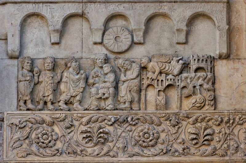 Fidenza, duomo (Cathedral of San Donnino), sculptures between the main portal and the left one: “Adoration of the Magi and Joseph’s dream”. Work by Benedetto Antelami and workshop.
On the left, the three Magi pay homage to the blessing Child sitting on the lap of the Mother in majesty; next to the Madonna, St. Joseph, sitting in profile, sleeping.
On the right, in a landscape characterized by the presence of buildings, an angel flies to Joseph to speak to him in a dream. On the far right, in the lower part, two doves in a garden drink from a fountain resting on flowering clods that represent the land of Egypt.
On the two sandstone panels placed above the other two, a frame with five hanging arches is sculpted and under the central one – above the heads of the Madonna and Joseph – a disk in the shape of a millstone symbolizes the star that showed the way to the Magi.
On the arches, the following inscriptions are engraved in capital letters: CASPAR . BALTA SAR . MELCHIOR MARIA . IHVS...