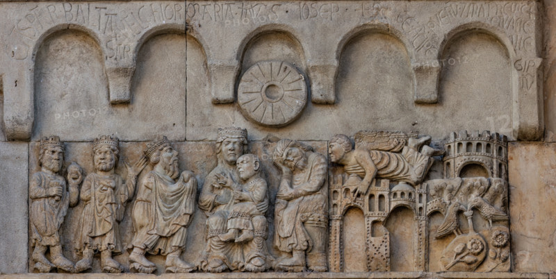 Fidenza, duomo (Cathedral of San Donnino), sculptures between the main portal and the left one: “Adoration of the Magi and Joseph’s dream”. Work by Benedetto Antelami and workshop.
On the left, the three Magi pay homage to the blessing Child sitting on the lap of the Mother in majesty; next to the Madonna, St. Joseph, sitting in profile, sleeping.
On the right, in a landscape characterized by the presence of buildings, an angel flies to Joseph to speak to him in a dream. On the far right, in the lower part, two doves in a garden drink from a fountain resting on flowering clods that represent the land of Egypt.
On the two sandstone panels placed above the other two, a frame with five hanging arches is sculpted and under the central one – above the heads of the Madonna and Joseph – a disk in the shape of a millstone symbolizes the star that showed the way to the Magi.
On the arches, the following inscriptions are engraved in capital letters: CASPAR . BALTA SAR . MELCHIOR MARIA . IHVS...