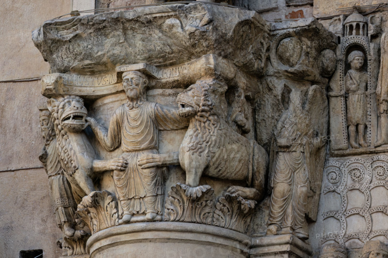 Fidenza, duomo (Cattedrale di San Donnino), sculture tra il portale maggiore e quello di sinistra. 
Particolare del semicapitello con “Daniele nella fossa dei leoni”. Il semicapitello figurato che raffigura nella parte frontale la scena biblica – di simbologia cristologica – del profeta Daniele che gettato nella fossa è attorniato dai leoni ammansiti. Sua lato destro del semicapitello è raffigurato il profeta Abacuc con il cibo per Daniele e sul lato sinistro l’angelo che lo portò (Dn 14,33-38). Sull’abaco del semicapitello è incisa a lettere capitali l’iscrizione: […]C DANIEL IVSTUS INLA EST CV LEONV[M] ([Abacu]c . Daniel iustus in lacu est cum leonum).
Opera di Benedetto Antelami e bottega.

[ENG]
Fidenza, cathedral (Cathedral of San Donnino), sculptures between the main portal and the left one:
Detail of the capital with “Daniel in the lions’ den”. The figurative semi-capital that depicts on the front the biblical scene – of Christological symbolism – of the prophet Daniel who...