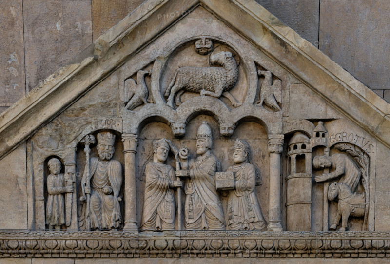 Fidenza, duomo (Cattedrale di San Donnino), portale sinistro, timpano. Nel frontone si leggono, da sinistra, tre scene: L'imperatore Carlo Magno, con un armigero alla sua destra;
Papa Adriano II che porge all'arciprete di San Donnino la mitria e il pastorale (simboli della dignità episcopale); il miracolo dell'ammalato guarito (miracolo compiuto da San Donnino).
Nell’acroterio sul timpano, é raffigurato un personaggio, probabilmente Oberto II Pallavicino condottiero posto da Federico II di Svevia alla guida della città dal 1249 al 1268.

Opera di Benedetto Antelami e bottega.

[ENG]
Fidenza, duomo (Cathedral of San Donnino), left portal, tympanum. In the pediment, from the left, three scenes can be read: Emperor Charlemagne, with a man-at-arms to his right;
Pope Adrian II who offers the archpriest of San Donnino the mitre and the pastoral staff (symbols of episcopal dignity); the miracle of the healed sick man (miracle performed by San Donnino).
In the acroterion on the tympan...