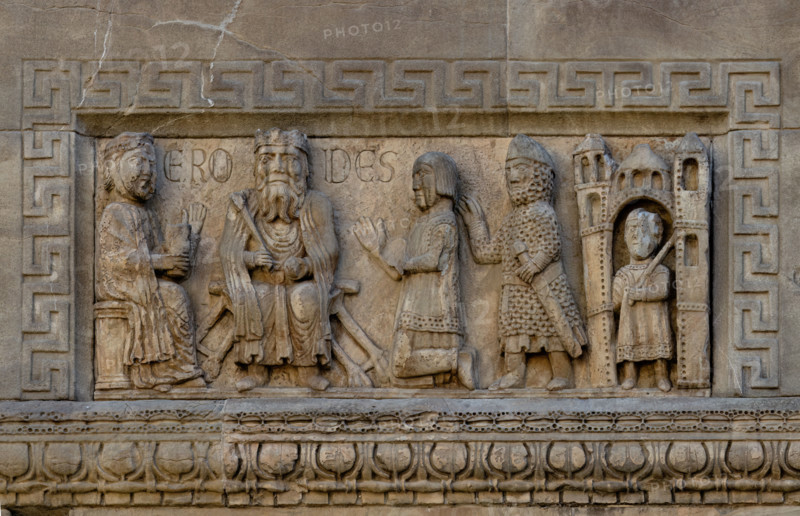 Fidenza, duomo (Cathedral of San Donnino), façade, sculptures between the left portal and the main portal: 
862 / 5.000
“King Herod”.
The slab depicts Herod, king of Israel from 37 to 4 BC – sitting on a faldstool, in medieval clothes and with the royal insignia.
To his right, seated in profile, a female figure (the queen?) holding a cylindrical container in her left hand.
To the left of the sovereign, a kneeling nobleman presented by a soldier armed with a sword and dressed in a medieval helmet and coat of mail. To the far right, under a building, a man armed with a sword and dressed in an ornate tunic. On the slab, to the right and left of the king’s head, the inscription ERODES is engraved.
It is not unlikely that the scene represents the moment in which the sovereign orders the Massacre of the Innocents (Mt 2:16-18), which would be coherently connected with the Adoration of the Magi, Joseph’s Dream and the Flight of the Three Kings, depicted in other bas-reliefs on the façade...