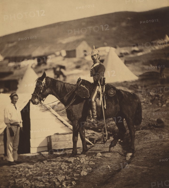 Full-length Portrait of British 5th Dragoon Guards Captain in Uniform, Seated on Horse with another Man Standing at Head of Horse, Tent and Huts in Background, Crimean War, Crimea, Ukraine, by Roger Fenton, 1855