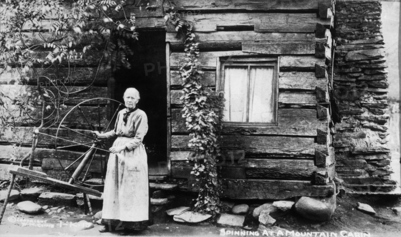 Woman with Spinning Wheel Beside Log Cabin, circa 1910