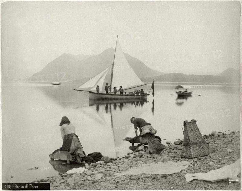 Two Women Doing Laundry at Shore of Lake Maggiore with Sailboat and Sasso di Ferro Mountain in Background, Lombardy, Italy, circa 1880