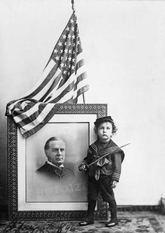 Boy in Sailor Uniform holding Sword in front of flag-draped, head-and-shoulders portrait of William McKinley, 1896