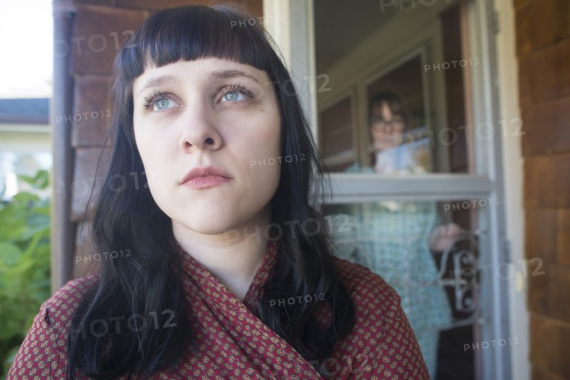 Head and Shoulders Portrait of Contemplative Young Adult Woman with Mid-Adult Woman in Background Looking at her through Front Door