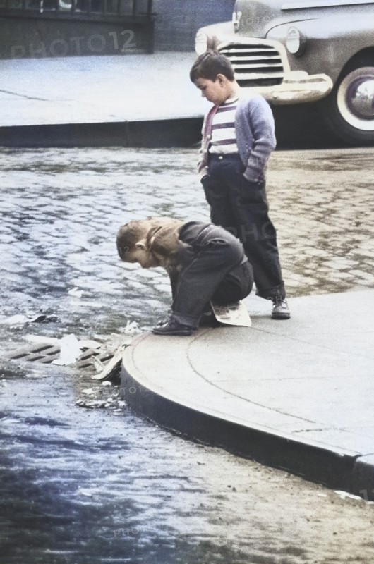 Two Young Boys on Street Corner looking in Storm Drain