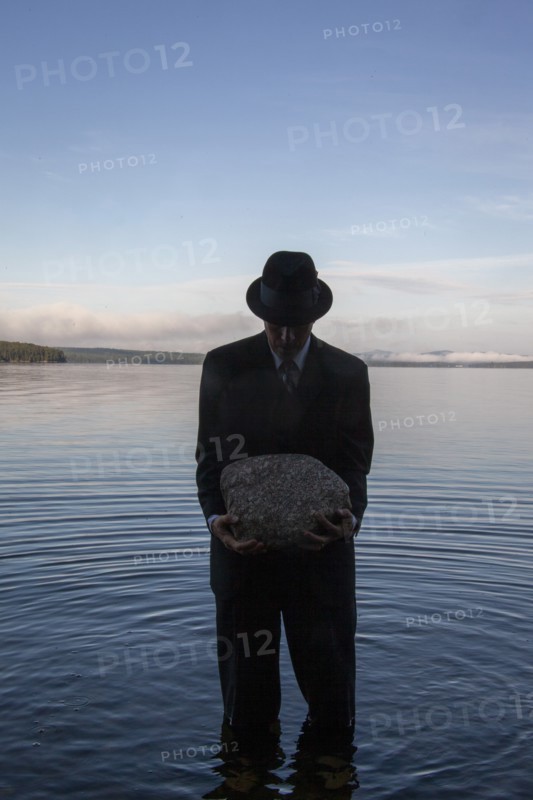 Man in suit and hat holding boulder while standing in calm lake at sunset