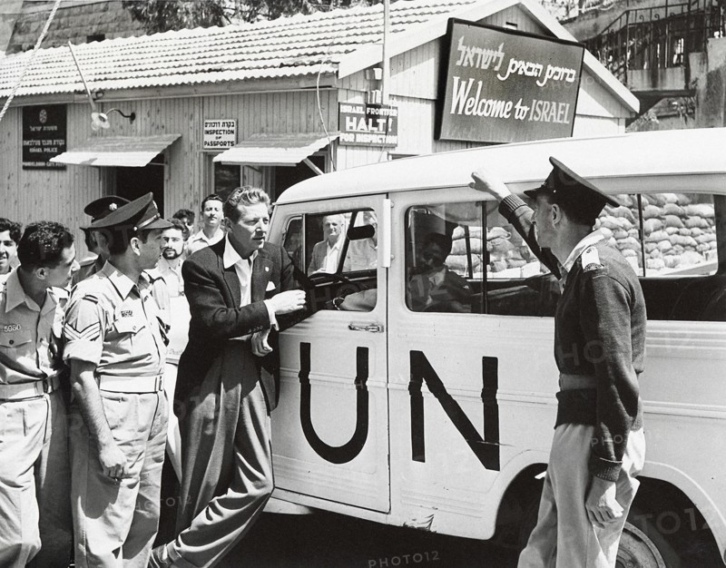 Danny Kaye leaning against UN vehicle near sign saying "Welcome to Israel"