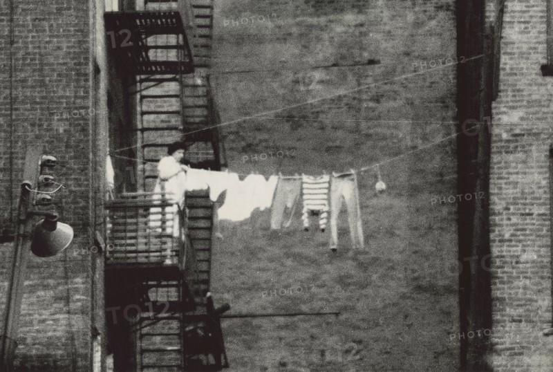 Woman standing on fire escape hanging clothes to dry on clothes line running to neighboring building