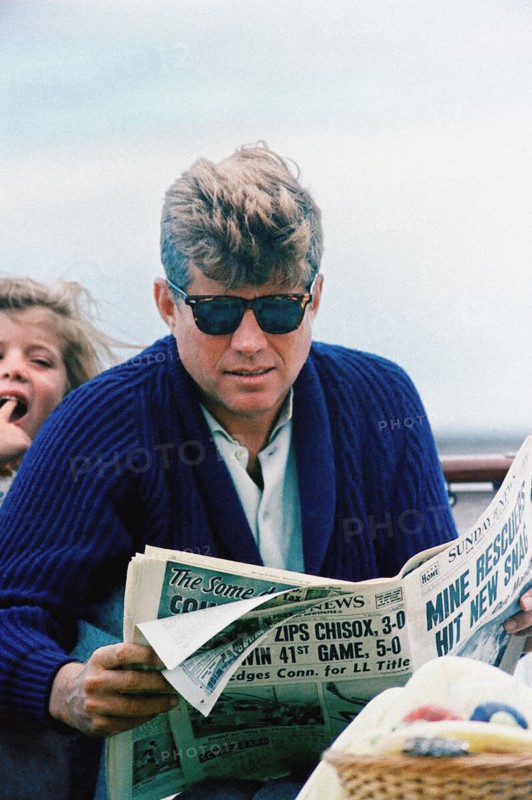 U.S. President John F. Kennedy reading newspaper with  his daughter, Caroline Kennedy in background, aboard the Presidential yacht, “Honey Fitz,” off the coast of Hyannis Port, Massachusetts, USA, Cecil Stoughton, White House Photographs, August 25, 1963