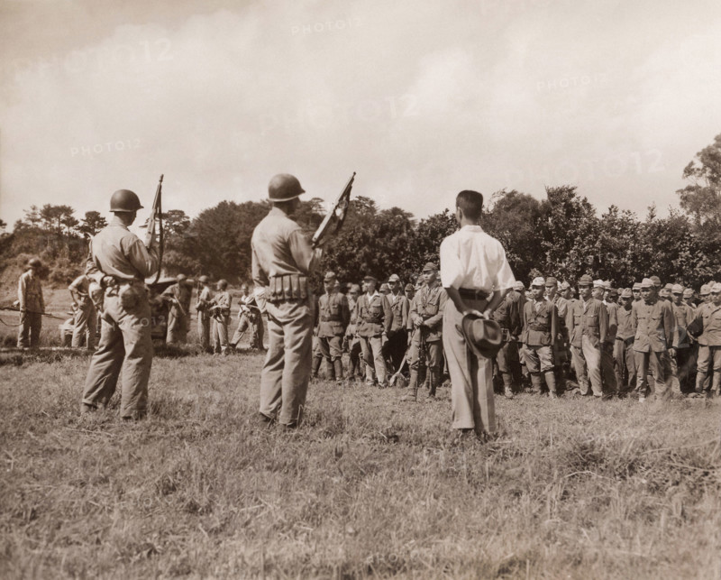 Japanese soldiers line up for formal surrender ceremonies as U.S. Marines with tommy guns and fixed bayonets stand guard, Motobu Peninsula, Okinawa, W. Huntington, U.S. Marine Corps, September 2, 1945