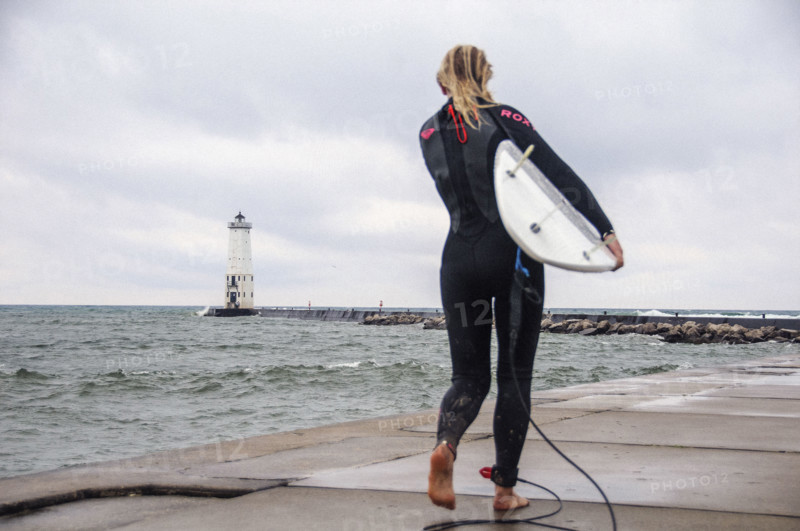 Rear view of young adult woman in wetsuit standing with surf board on Frankfort North lighthouse and pier, Lake Michigan, Frankfort, Michigan, USA