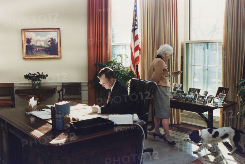 U.S. President George H.W. Bush working at his desk in the Oval Office as U.S. First Lady Barbara Bush looks at photographs on the table behind desk, White House, Washington, D.C., USA, Susan Biddle, White House photographer, January 27, 1989