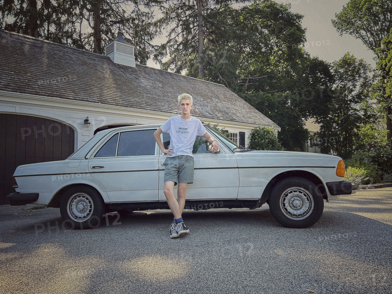 Young adult man with bleached hair leaning against vintage Mercedes car in driveway