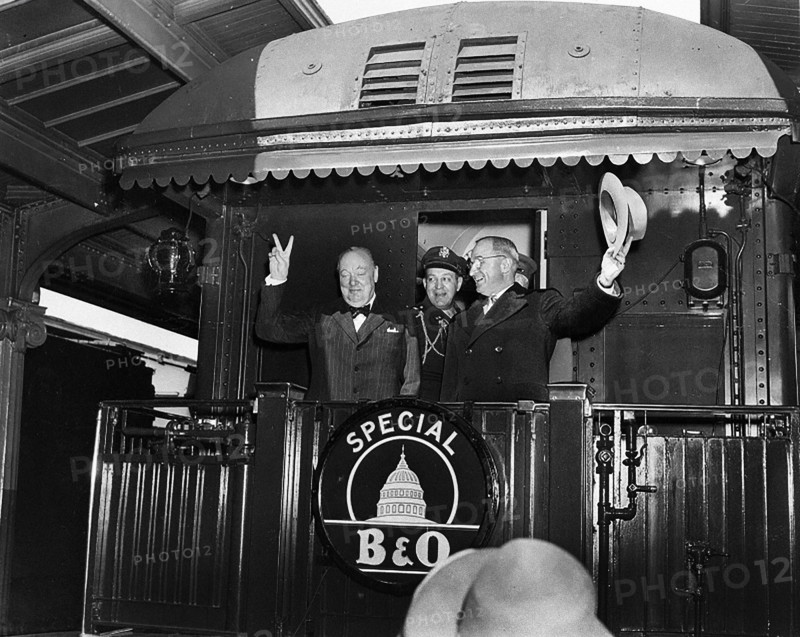 U.S. President Harry S. Truman (right) waving his hat standing to right of former British Prime Minister Winston Churchill flashing his famous "V for Victory" sign from rear platform of special Baltimore & Ohio train, en route to Fulton, Missouri for Churchill's "Iron Curtain" speech, as President's Military Aide, General Harry Vaughan, looks on, Washington, D.C., USA, Abbie Rowe, National Parks Service, Harry S. Truman Library, March 4, 1946