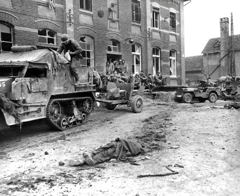 Members of Company G, 2nd Battalion, 16th regiment, 1st Division, U.S. First Army with military half-track hybrid vehicle waiting for orders to move on, as dead German soldier lays on ground in foreground, Vetteweiss, Germany, U.S, Army Signal Corps, February 28, 1945