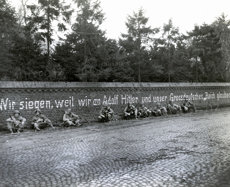 Members of 16th regiment, 1st Division, U.S. First Army, sitting under German slogan "Wir Siegen, weil wir an Adolf Hitler und unser Grossdeutches Reich Glauben", English translation, "We triumph because we believe in Adolf Hitler and our Greater German Reich", Vetteweiss, Germany, U.S, Army Signal Corps, February 28, 1945