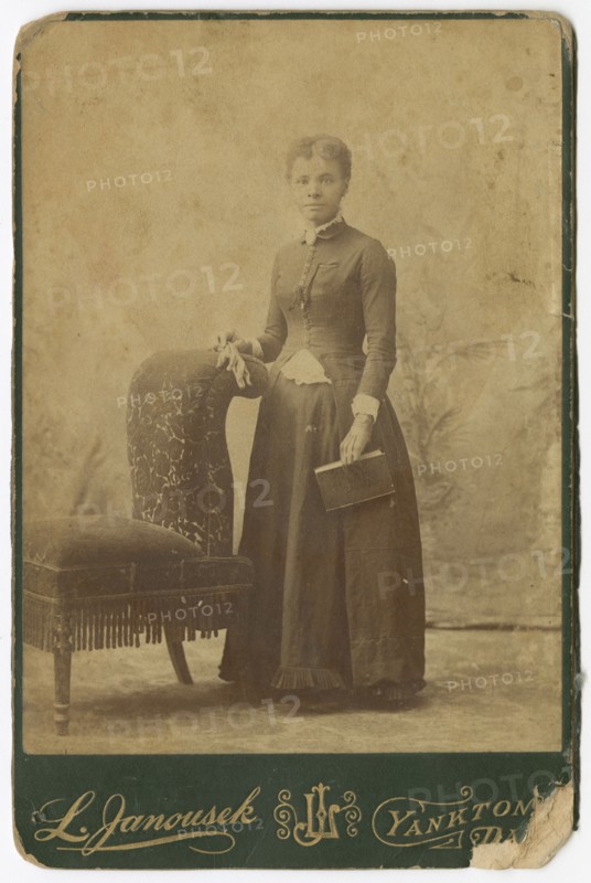 Photograph of a woman standing next to a chair and holding a book, late 19th century. Creator: Louis Janousek.