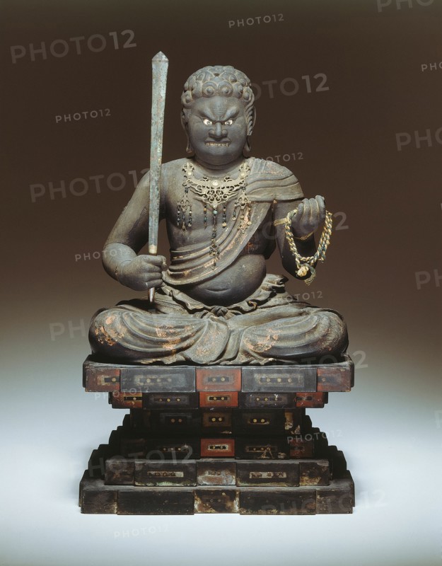 Fudo Myo-o, 13th century. A wooden sculpture of an angry looking diety, holding a sword in his right hand and ope in his left, sitting cross-legged on an stylized rock formation