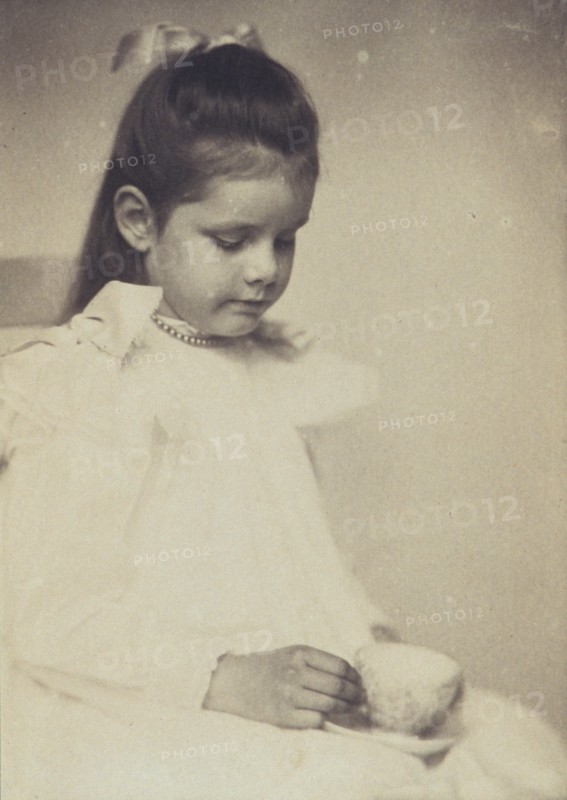 Young girl in white dress, seated holding teacup and saucer, c1900.