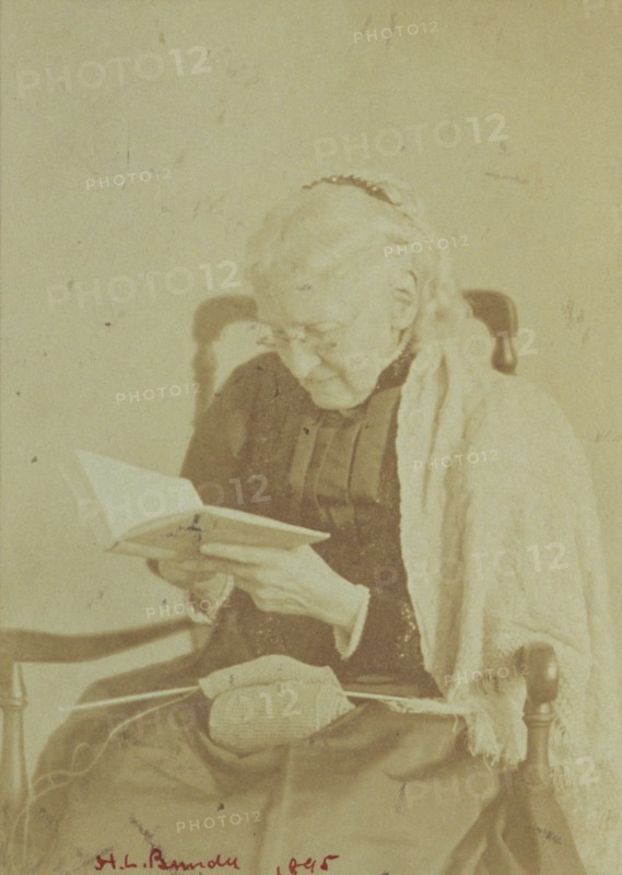 Elderly woman seated and reading a book with knitting in her lap, 1895.