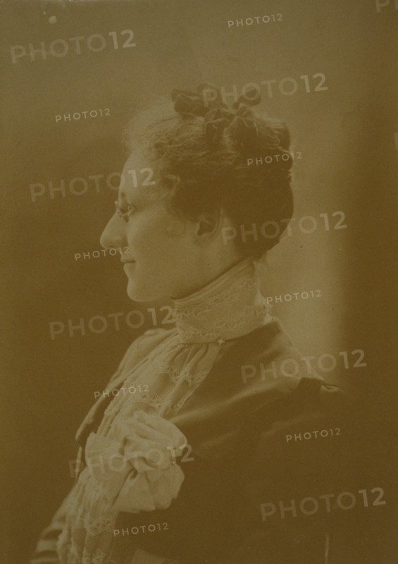 Studio portrait of young woman in white dress posed as if holding flowering tree branch, c1900.