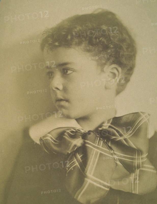 Head-and-shoulders portrait of a young boy with a large plaid bow at the neck of his jacket, facing left, c1900.